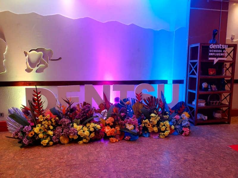 Cover of the Dentsu School of Influence album: a white DENTSU letter installation banked with tropical florals under purple-and-blue lighting