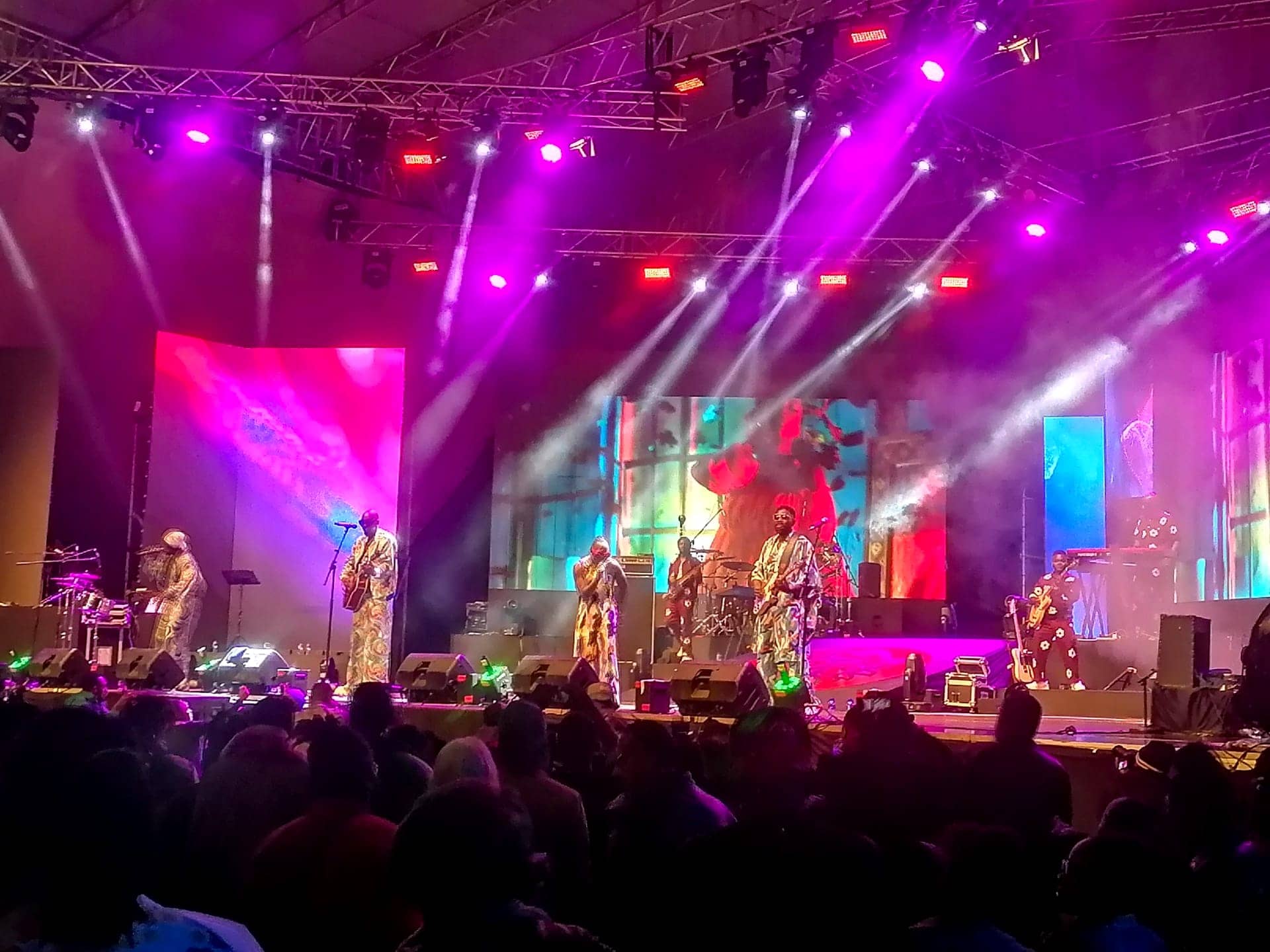 A band in patterned outfits performing on a large festival stage under magenta and white beam lighting, with a silhouetted crowd in front