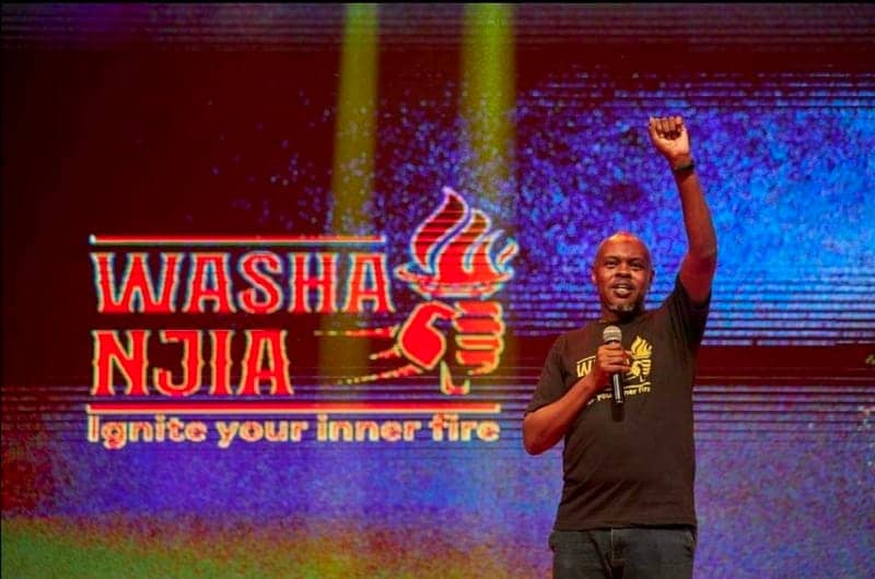 A speaker raises a fist on stage in front of a glowing 'Washa Njia — Ignite your inner fire' LED wall