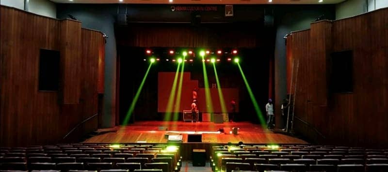 Green moving-head beams fan across an empty theatre stage at the Kenya Cultural Centre, rows of seats in the foreground